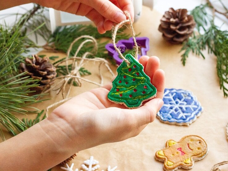 Selbstgebastelter Christbaumanhänger aus Salzteig in Kinderhand, dahinter bunte Kekse, Tannenzweige und ein violetter Ausstecher.