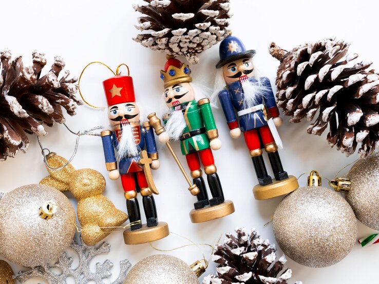 Drei bunte Nussknacker mit weißem Bart stehen zwischen glitzernden Christbaumkugeln und schneebedeckten Tannenzapfen.