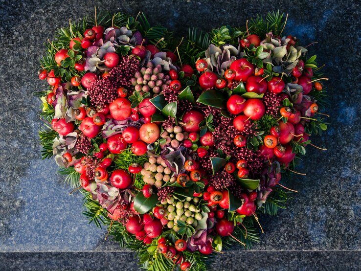 Herzförmiges Grabgesteck für Totensonntag  aus roten Beeren, Hortensien, Blättern und Tannenzweigen auf dunkelgrauem Naturstein.