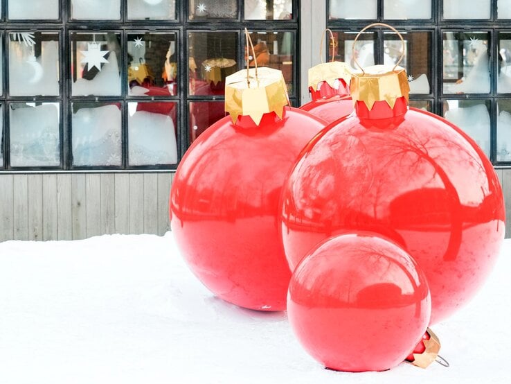 Vier übergroße rote Christbaumkugeln mit goldenen Aufhängern stehen dekorativ im Schnee vor einem Fenster mit Eisblumen.