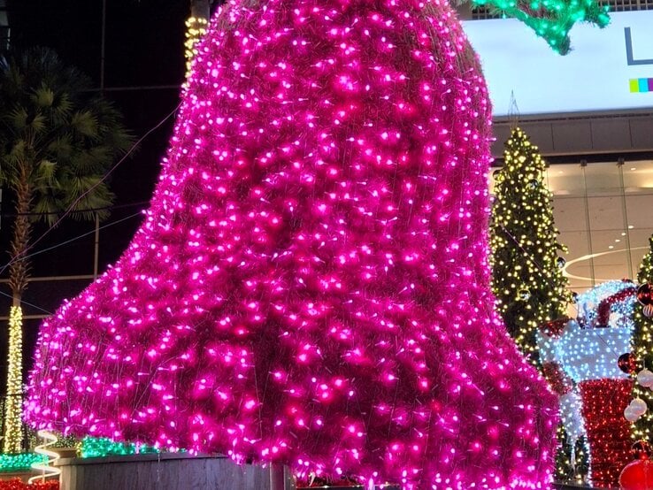 Riesige, pink leuchtende Weihnachtsglocke mit grünem Lichtschleifen über einem Wasserbecken vor festlich geschmücktem Gebäude.