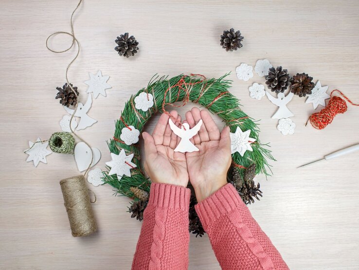 Zwei Hände in rosa Strick halten einen weißen Engel aus Salzteig über einem festlich geschmückten Adventskranz.