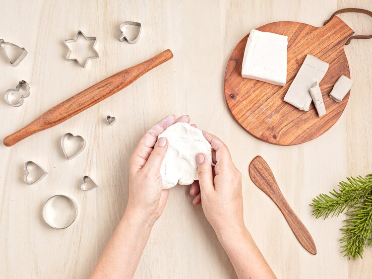 Zwei Hände formen helle Modelliermasse auf hellem Holztisch, umgeben von Ausstechformen, Holzrolle und Tannenast.