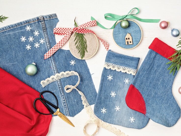 Selbstgemachte Weihnachtsdeko aus Jeans, Stickrahmen und Spitze liegt auf weißem Holz mit Kugeln und Tannenzweigen.