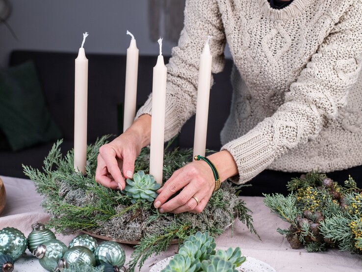 Eine Frau mit beigem Strickpullover dekoriert einen Adventskranz mit Sukkulenten und vier weißen Stabkerzen auf Moosgrund.