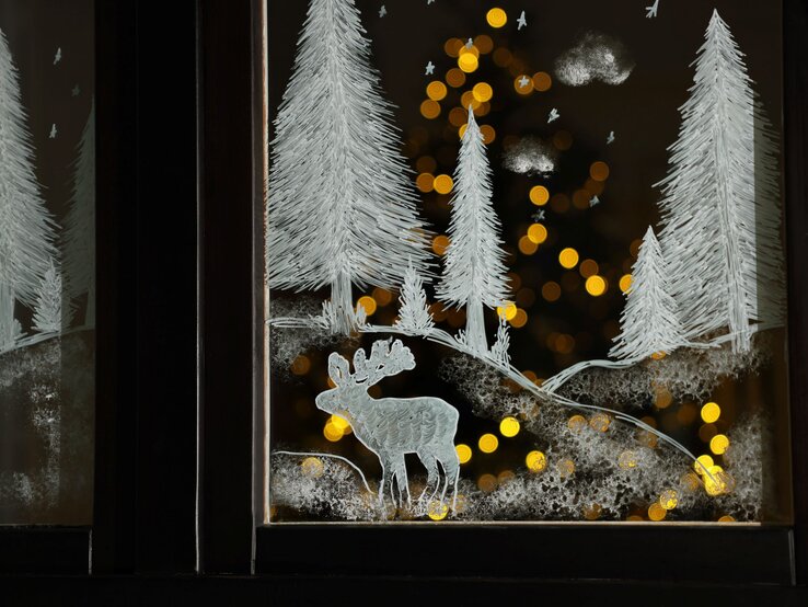 Fensterdeko aus Buttermilch und Kefir: Auf ein Fenster gemaltes Winterbild mit Tannen, Rentier und Schneelandschaft, dahinter warme, goldene Lichter verschwommen.