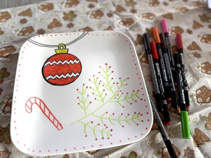 Weißer Teller mit handgemalten Weihnachtsmotiven wie Kugel, Zuckerstange und Zweig, daneben bunte Edding-Stifte.