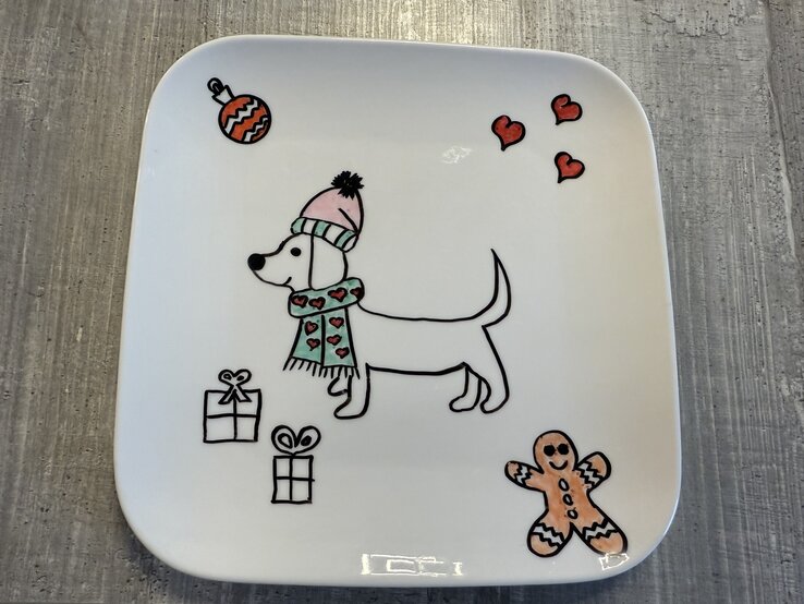 Weihnachtsteller bemalen: Zeichnung eines Dackels mit rosa Mütze und grünem Schal, umgeben von Geschenken, Herzen, Kugel und Lebkuchenmann. | © bildderfrau.de/Madeleine Liedke 