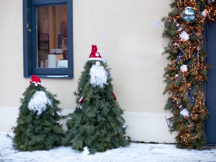 Tannenförmige Weihnachtsfiguren mit Lockenbärten und Zipfelmützen stehen im Schnee neben beleuchtetem Türkranz.
