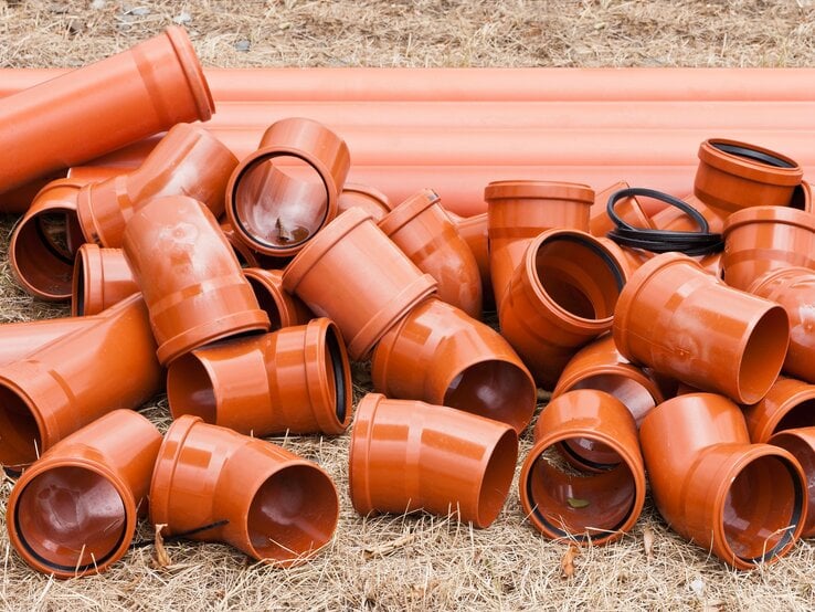 Mehrere orange Abflussrohre und HT-Rohre aus Kunststoff liegen ungeordnet auf trockenem, braunem Grasboden.