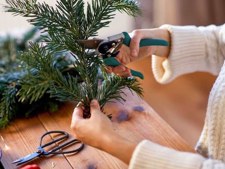Eine Person in cremefarbenem Strickpullover schneidet mit Gartenschere frische Tannenzweige auf einem Holztisch.