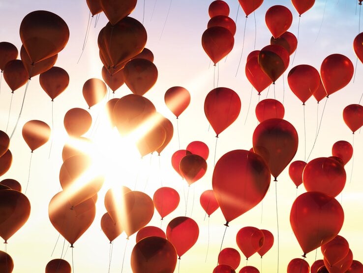 Leuchtend rote Luftballons schweben vor strahlender Sonne in den pastellfarbenen Himmel.