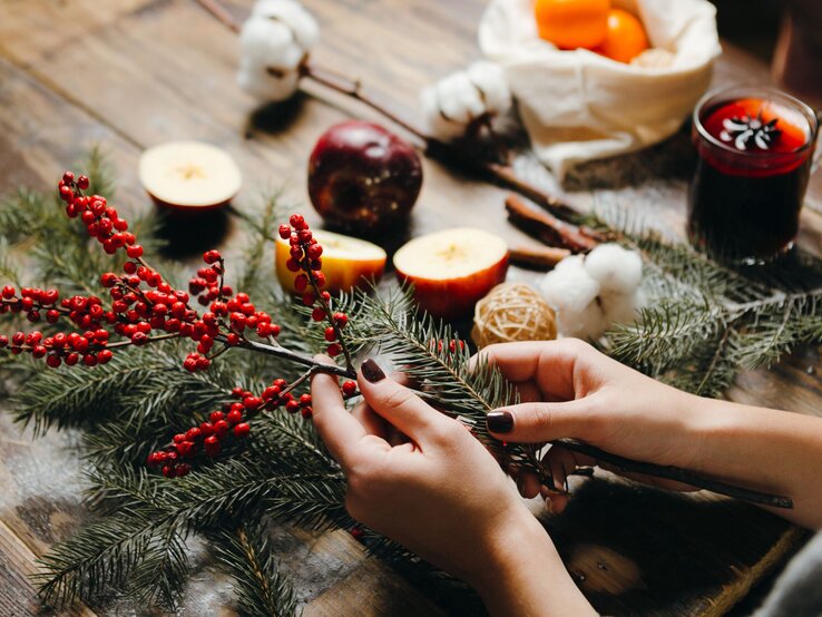 Zwei Hände mit dunkel lackierten Nägeln arrangieren Tannenzweige, Thuja und rote Beeren auf einem rustikalen Holztisch.