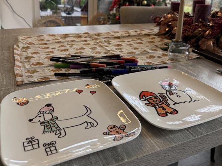 Zwei bemalte Weihnachtsteller mit Hund und Lebkuchenmann liegen auf einem Tisch vor festlich geschmücktem Weihnachtsbaum.