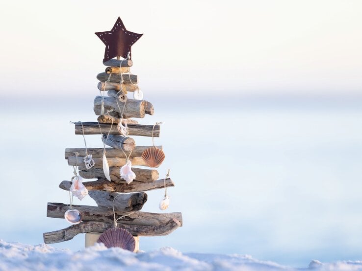 Aus Treibholz und Muscheln gebastelter Weihnachtsbaum mit Sternspitze steht auf hellem Sand vor sanftem Meereshorizont.