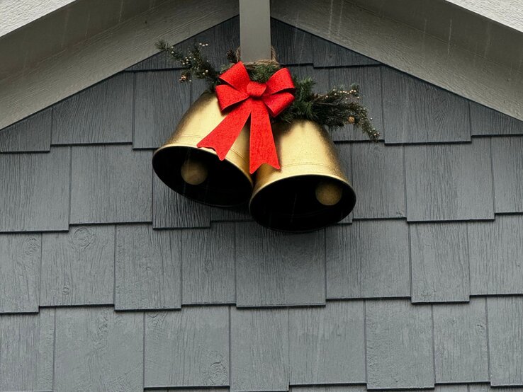 Zwei goldene Weihnachtsglocken mit roter Schleife und Tannenzweig hängen dekorativ an einer grau gestrichenen Hausfassade.