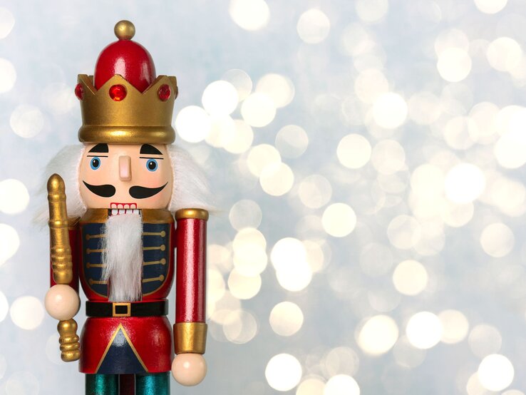 Bunt bemalter Nussknacker mit roter Krone und weißem Bart steht vor unscharf leuchtendem, winterlichem Hintergrund.