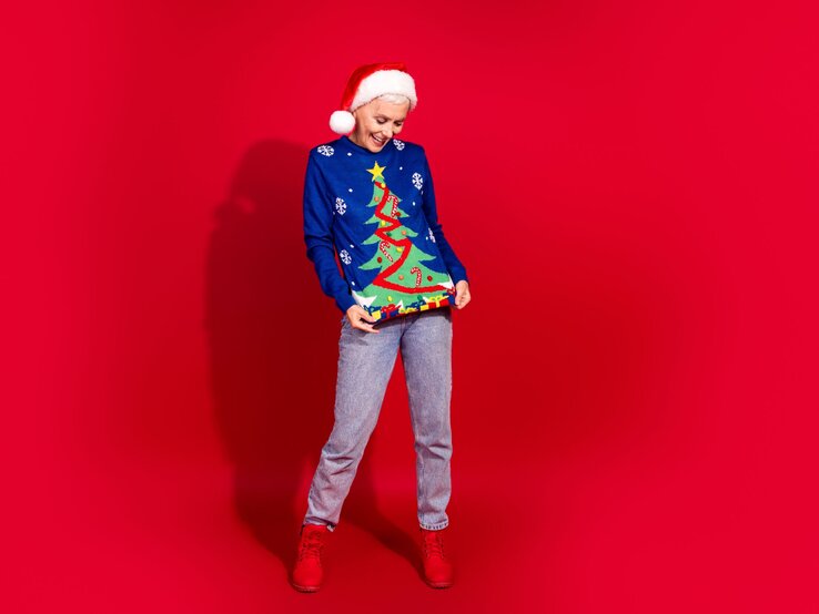 Lachende ältere Frau mit Weihnachtsmütze zeigt auf ihren blauen Weihnachtspullover mit Tannenbaummotiv vor knallrotem Hintergrund.