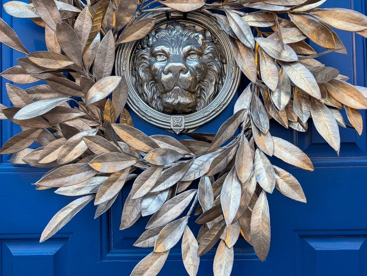 Ein kunstvoller Kranz aus goldenen Lorbeerblättern umrahmt einen Löwenkopf an einer kräftig blauen Holztür.