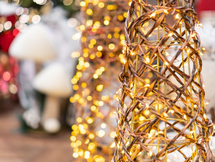 Geflochtener Drahtkegel mit warmweißen Lichterketten vor verschwommenem Weihnachtsdekor in Rot und Weiß.