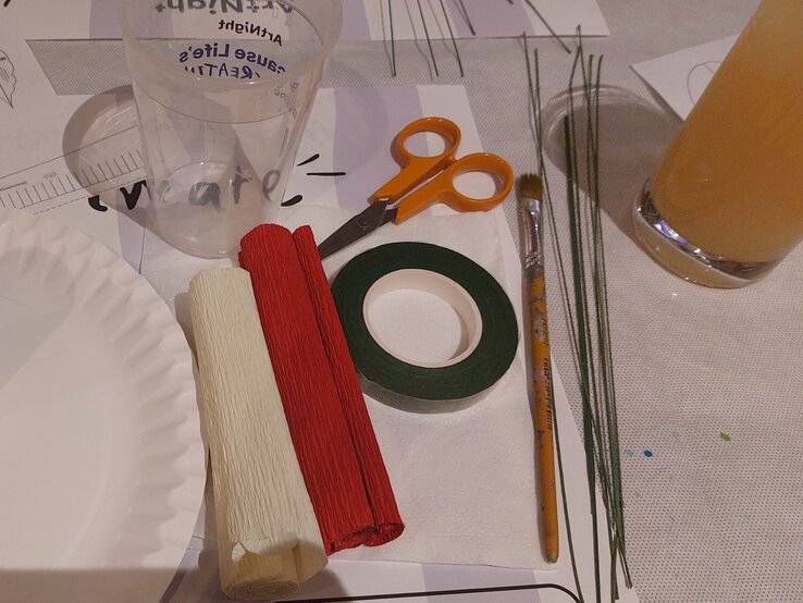 Bastelmaterialien auf einem Tisch: weißer Teller, Farbkrepppapier, Schere, Pinsel, Draht, Klebeband, leeres Plastikglas und orangensaftgefülltes Glas. | © Linda Pötzsch