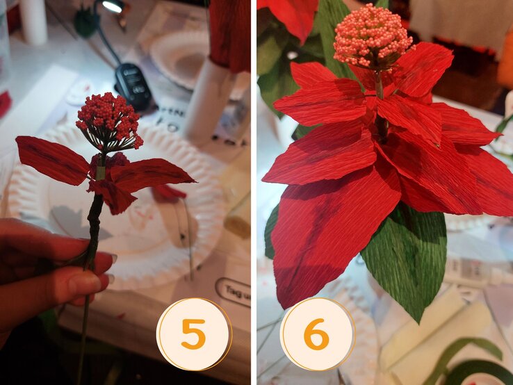 Halbfertige Krepppapierblüte mit roten Blättern und einer perlenbesetzten Mitte, gehalten von einer Hand vor einem Basteltisch mit Pappteller und Klebezubehör. | © Linda Pötzsch