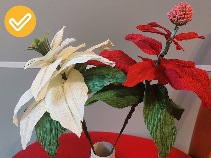 Zwei detailreiche Krepppapierblumen in Weiß und Rot mit grünen Blättern, kunstvoll arrangiert auf einem roten Tisch vor grauem Hintergrund. | © Linda Pötzsch