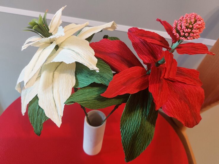 Zwei handgefertigte Krepppapierblumen, eine in Rot und eine in Weiß, mit grünen Blättern in eleganter Vase auf rotem Tisch vor grauem Hintergrund.