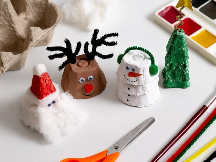 Vier weihnachtliche Figuren aus Eierkarton – Nikolaus, Rentier, Schneemann und Tannenbaum – stehen auf hellem Basteltisch.