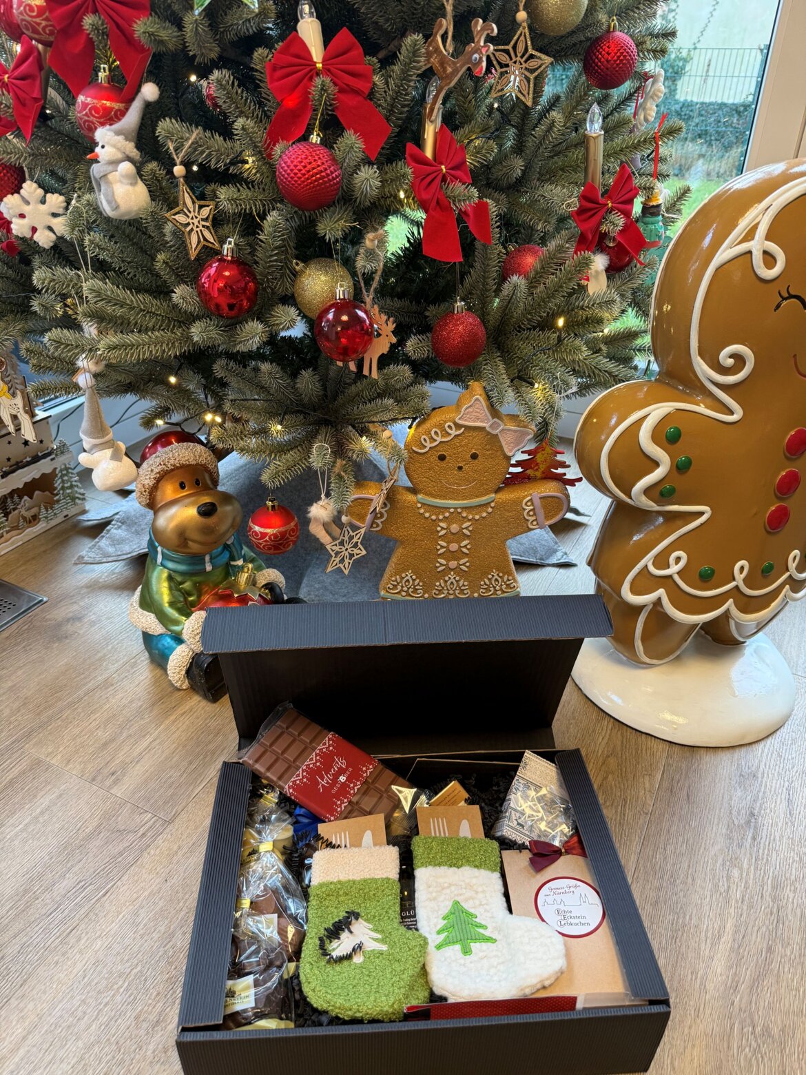 Geöffnete Geschenkbox mit Socken, Schokolade und Naschwerk liegt vor einem festlich geschmückten Weihnachtsbaum mit Dekofiguren. | © Madeleine Liedke 
