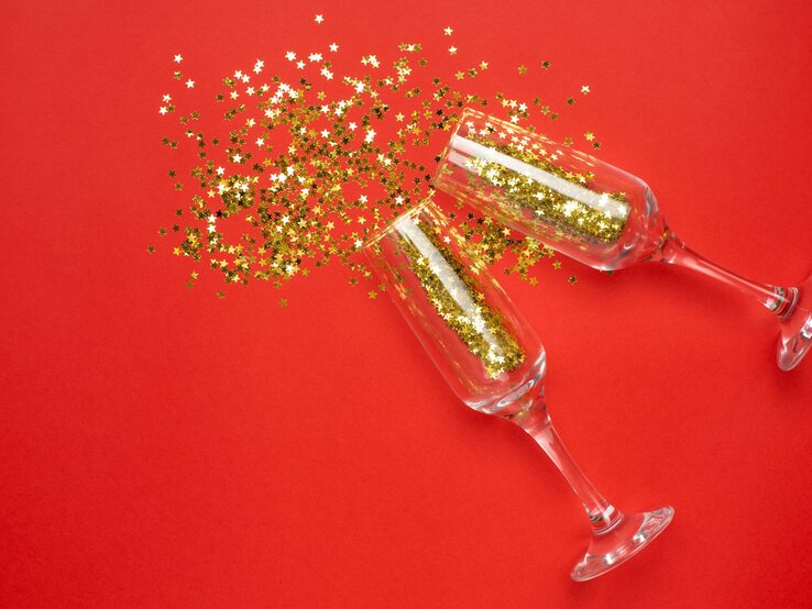 Zwei Sektgläser stoßen an, goldene Stern-Konfetti wirbeln sprühend über rotem Hintergrund durch die Luft.