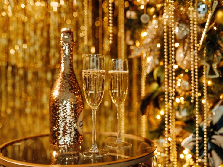 Zwei gefüllte Sektgläser und eine glitzernde Sekt-Flasche stehen vor goldenem Lametta und funkelndem Christbaumschmuck.