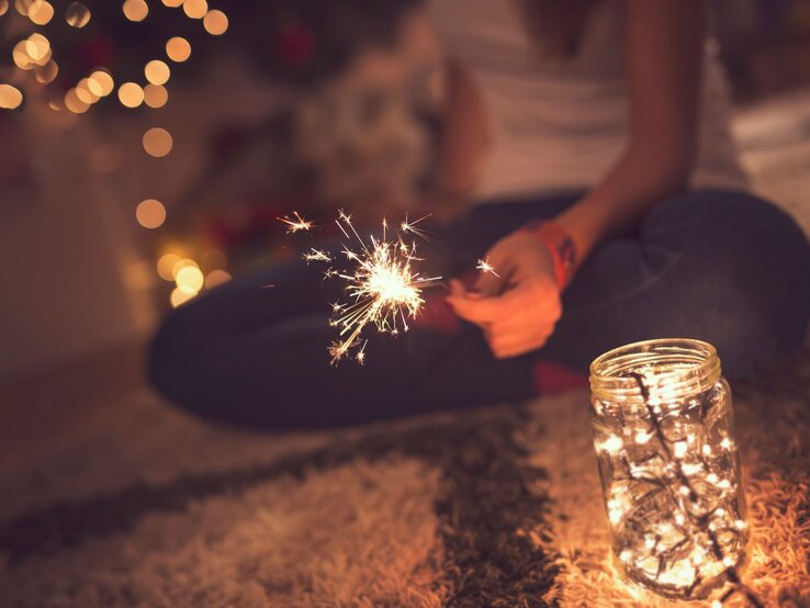 Junge Frau mit Wunderkerze sitzt im Schneidersitz auf Teppich, neben ihr ein Windlicht mit warm leuchtender Lichterkette.