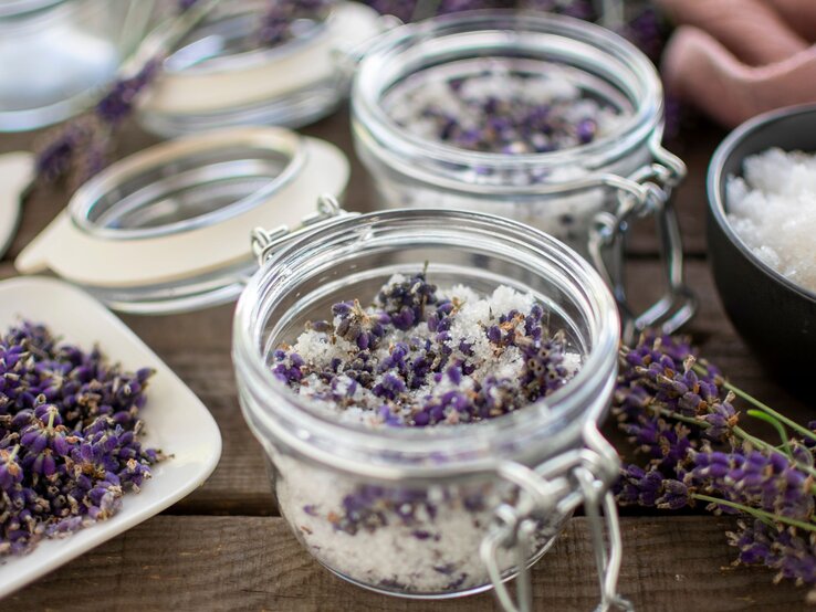 Ein Glas mit grobem Salz und getrocknetem Lavendel steht offen auf rustikalem Holz, daneben Blüten, Zucker und weitere Gläser.