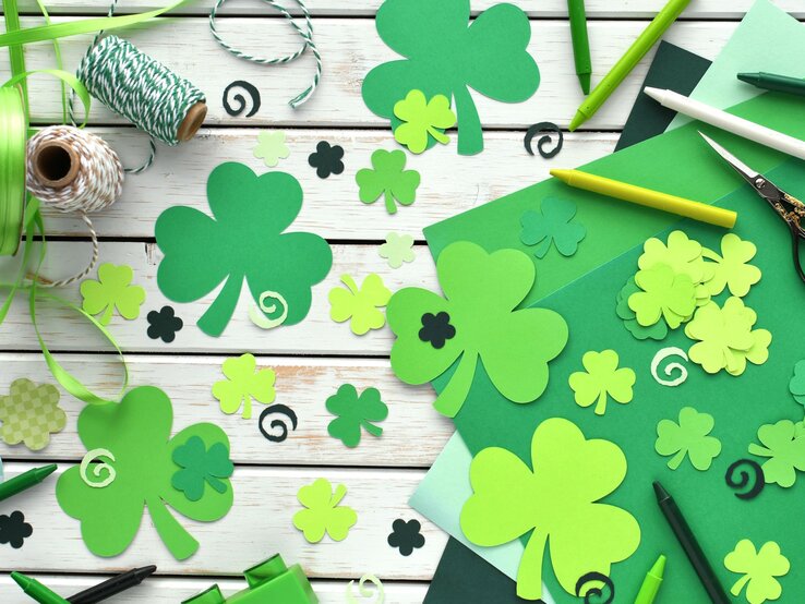 Bunte Bastelmaterialien und ausgeschnittene grüne Kleeblätter liegen auf weißem Holzuntergrund in fröhlichem Chaos.