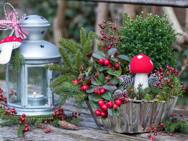 Silvster-Deko mit Filzfliegenpilz, roten Beeren und Tannenzweigen in einer Schale neben silberner Laterne mit Kerze.