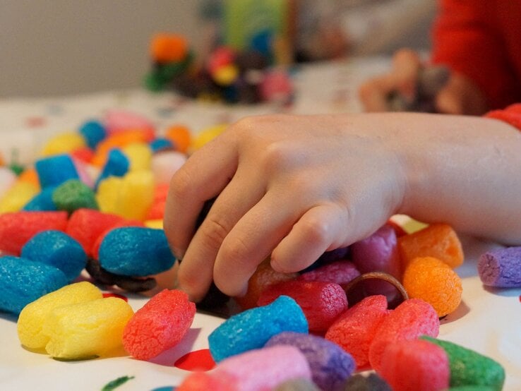 Kinderhand greift nach bunten Bastelchips aus Mais auf einem weißen Tisch mit roten Punkten im weichen Tageslicht.