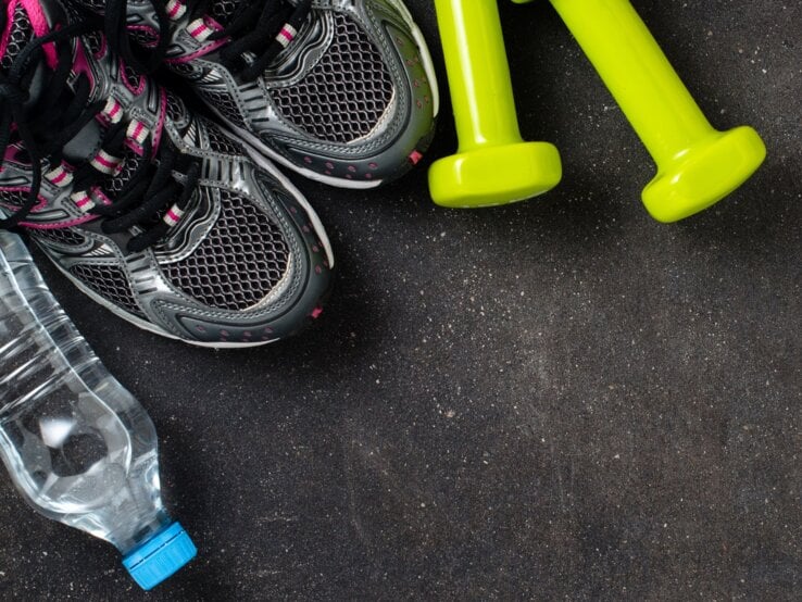 Zwei neongelbe Hanteln liegen neben grauen Sportschuhen mit pinken Details und einer Wasserflasche auf Asphaltboden.