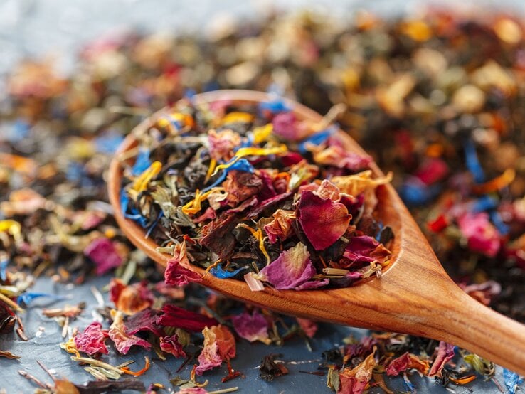 Bunte Blütenblätter und Teeblätter in kräftigen Farben auf einem Holzlöffel, lose verteilt auf dunklem Hintergrund.