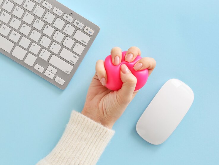 Eine Hand in cremeweißem Strickärmel drückt einen pinken Stressball neben Tastatur und Maus auf hellblauem Untergrund.