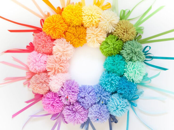 Regenbogenfarbener Pompon-Kranz aus weicher Wolle mit farbigen Geschenkbändern auf strahlend weißem Hintergrund arrangiert.