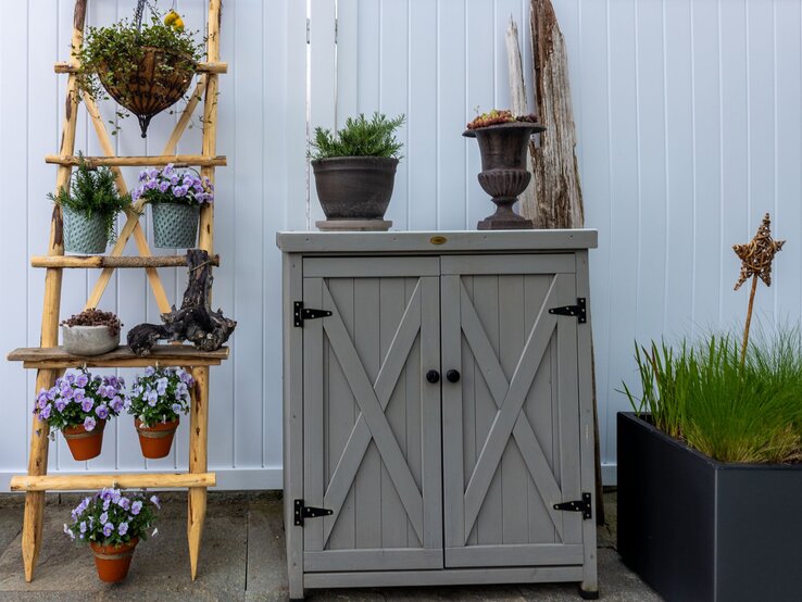 Blühende violette Blumen, Tontöpfe und Kräuter arrangiert auf Leiterregal neben rustikalem Gartenschrank aus Holz.