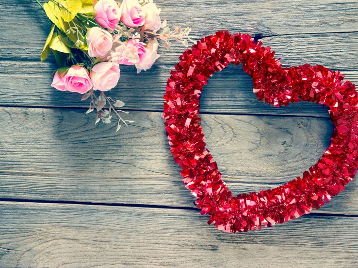 Ein Herz- Kranz aus roter Glitzergirlande liegt neben zarten rosa Rosen auf verwittertem Holztisch.