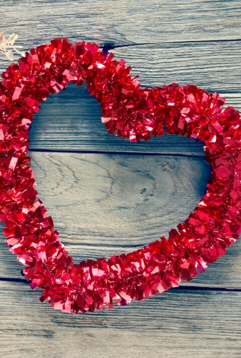 Ein Herz- Kranz aus roter Glitzergirlande liegt neben zarten rosa Rosen auf verwittertem Holztisch.