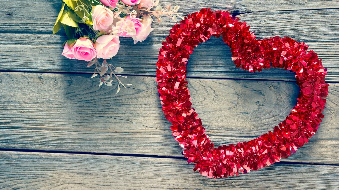 Ein Herz- Kranz aus roter Glitzergirlande liegt neben zarten rosa Rosen auf verwittertem Holztisch.
