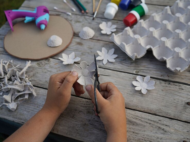 Kinderhände schneiden aus grauer Eierkartonpappe eine Blume, umgeben von Bastelmaterialien auf einem Holztisch.