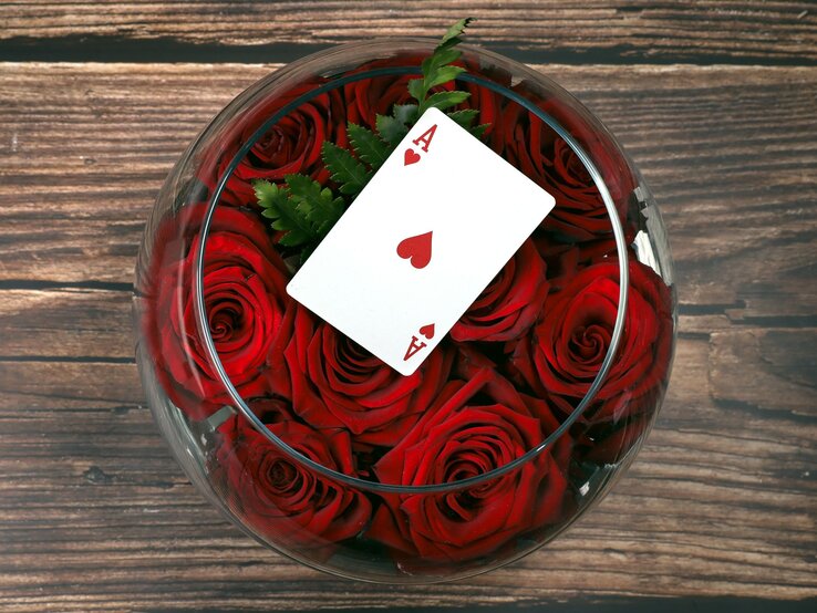 Eine Spielkarte mit Herz-Ass liegt auf roten Rosen in einer runden Glasvase vor rustikalem Holzhintergrund.