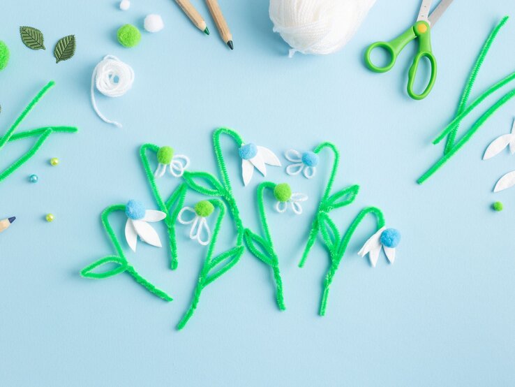 Auf hellblauem Untergrund liegen gebastelte Schneeglöckchen aus Eierkartons,Pfeifenreinigern, Pompons und Filzblättern in Frühlingsfarben.