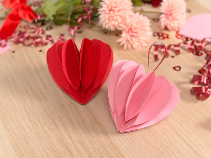 Rote und rosa Papierherzen stehen auf einem Holztisch, verziert mit pinken Schleifen, Blumen und Valentinstagsdekoration.