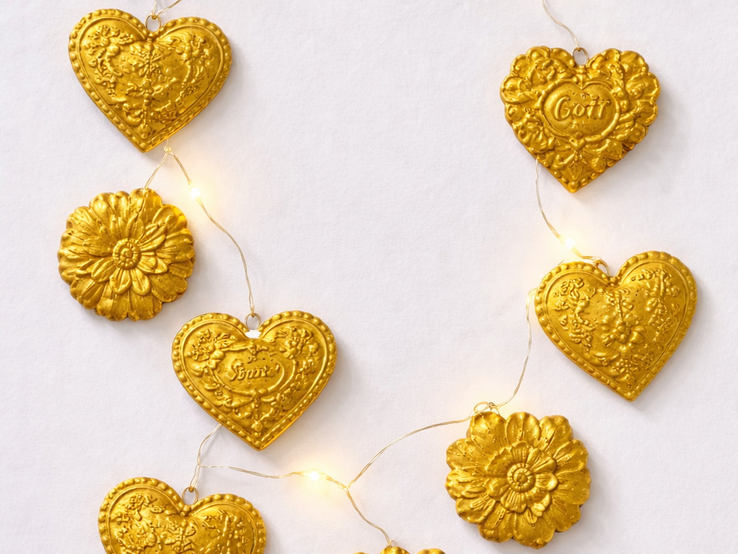 Goldene Herzen und Blumen mit Prägungen hängen an einer Lichterkette auf weißem Hintergrund in festlicher Anordnung.